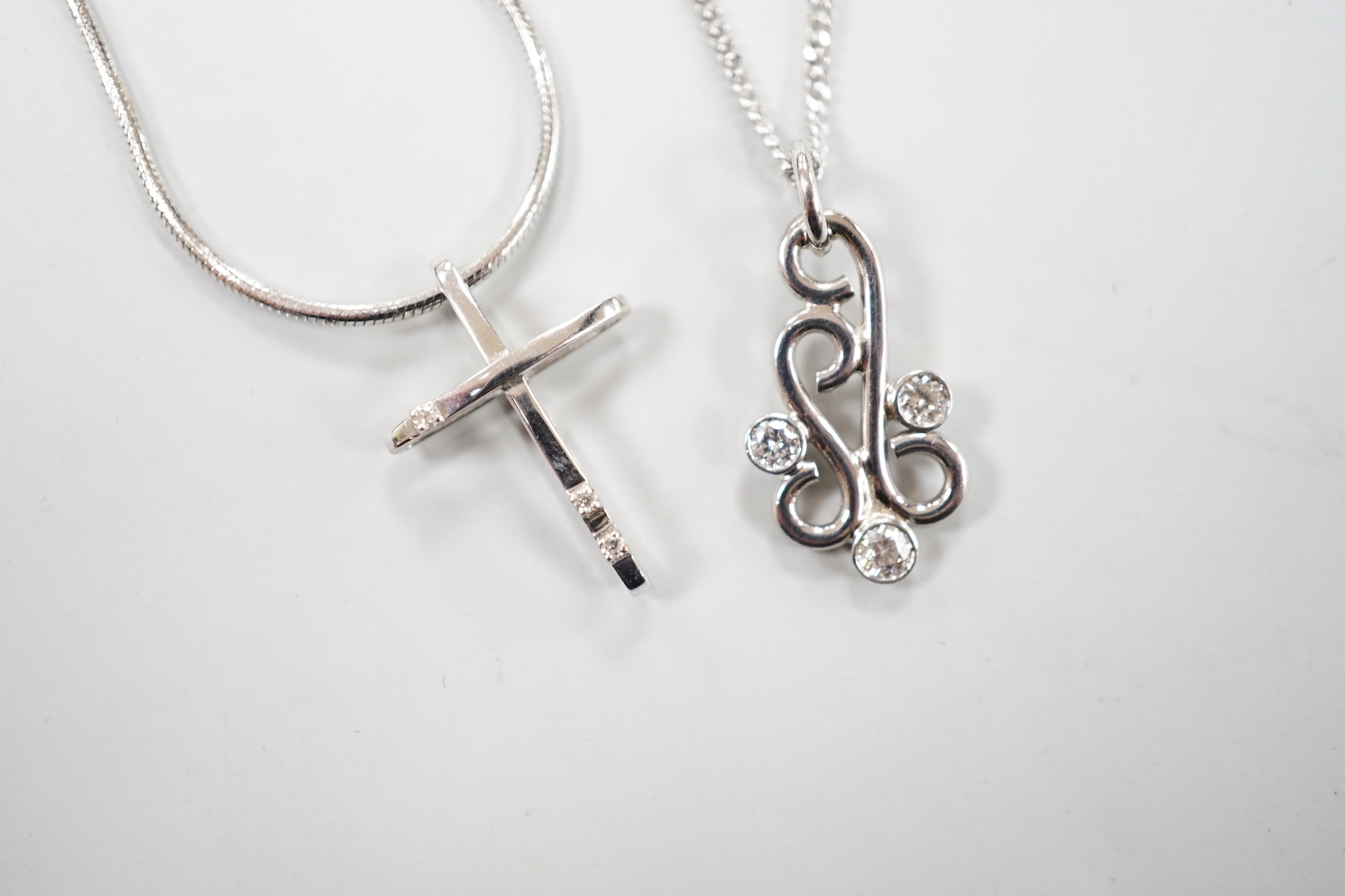 A modern 18ct white gold and three stone diamond set scroll pendant, 20mm, on an 18ct white gold chain, 39cm, gross weight 5.3 grams, together with a 585 white metal and diamond chip set cross pendant, on a 585 white met